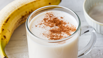 a glass of banana milk with a sprinkle of cinnamon on top and a banana next to it. Made in a flash with your Almond Cow.