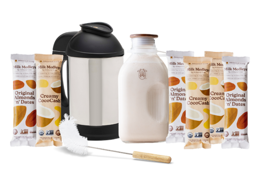 The Almond Cow Milk Maker with Glass Jug and Cleaning Brush surrounded by Almond 'n' Dates and Creamy CocoCash Milk Medleys